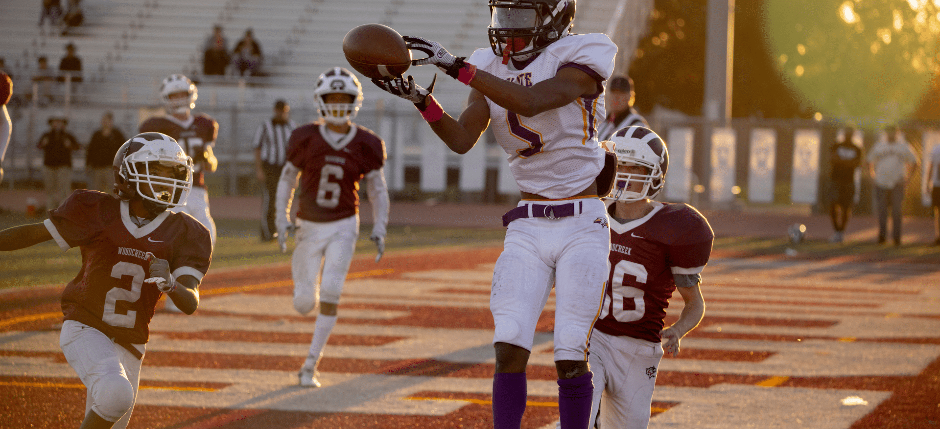 Football player in action catching a pass during a game, wearing protective gear including football pads, with two opposing players in the background on a sunlit field.