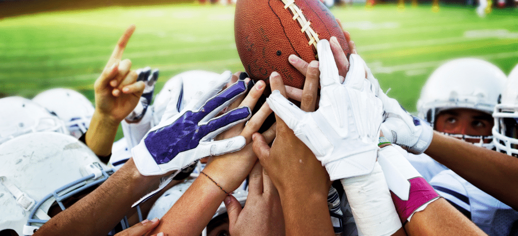 A group of football players' hands, wearing gloves, reaching together to hold a football, symbolizing teamwork, unity, and the spirit of high school football.